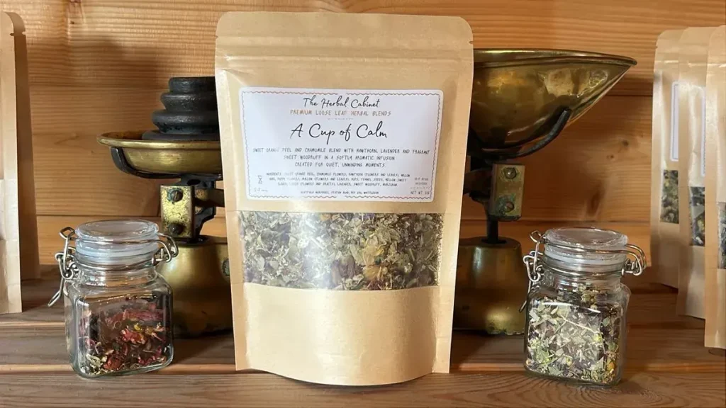 Kraft pouch of A Cup of Calm loose leaf herbal blend by The Herbal Cabinet, its window showing dried leaves with flecks of flower, beside brass scales and glass jars of herbs