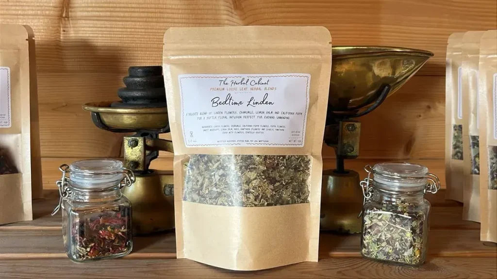Kraft pouch of Bedtime Linden loose leaf infusion by The Herbal Cabinet, its window showing pale green dried leaves and flowers, beside brass scales and glass jars of herbs