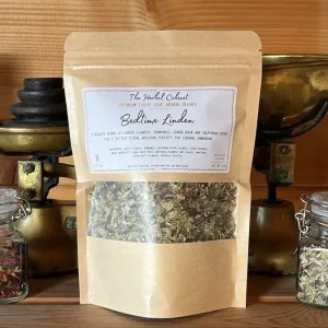 Kraft pouch of Bedtime Linden loose leaf infusion by The Herbal Cabinet, its window showing pale green dried leaves and flowers, beside brass scales and glass jars of herbs