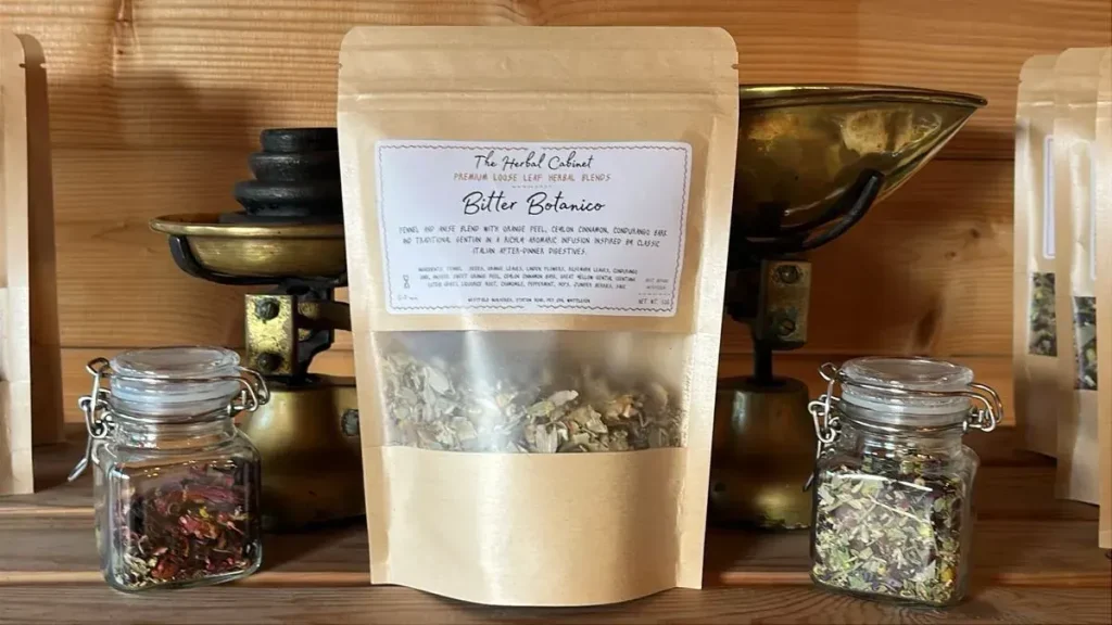 Kraft pouch of Bitter Botanico loose leaf infusion by The Herbal Cabinet, its window showing pale dried herbs and seeds, beside brass scales and glass jars of herbs