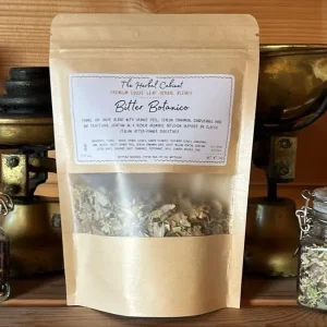 Kraft pouch of Bitter Botanico loose leaf infusion by The Herbal Cabinet, its window showing pale dried herbs and seeds, beside brass scales and glass jars of herbs
