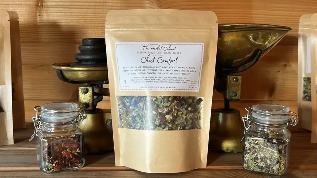 Kraft pouch of Chest Comfort loose leaf infusion by The Herbal Cabinet, its window showing green leaves with red petals and small yellow flower heads, beside brass scales and glass jars of herbs