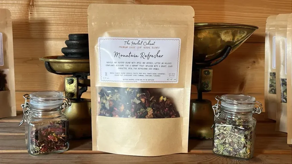 Kraft pouch of Mountain Refresher loose leaf infusion by The Herbal Cabinet, its window showing deep red hibiscus and fruit pieces, beside brass scales and glass jars of herbs