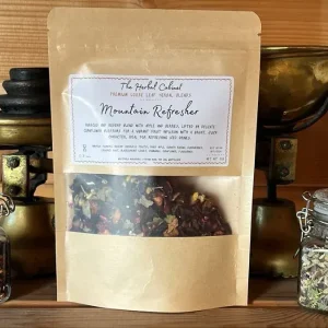 Kraft pouch of Mountain Refresher loose leaf infusion by The Herbal Cabinet, its window showing deep red hibiscus and fruit pieces, beside brass scales and glass jars of herbs