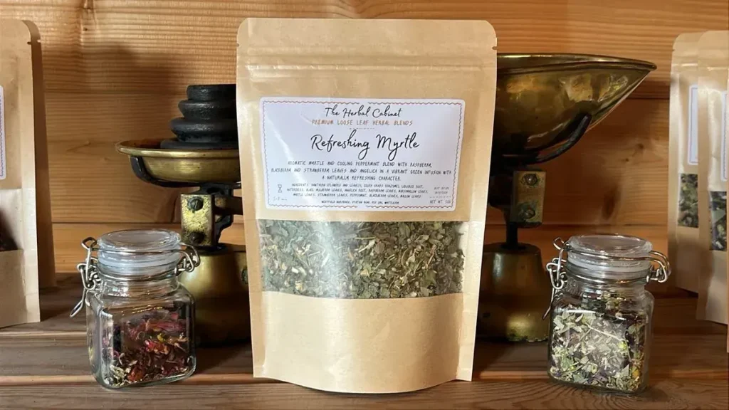 Kraft pouch of Refreshing Myrtle loose leaf infusion by The Herbal Cabinet, its window showing finely cut green leaves, beside brass scales and glass jars of herbs