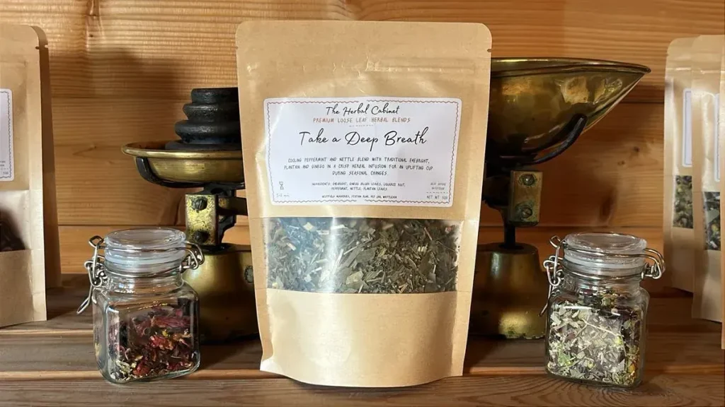 Kraft pouch of Take a Deep Breath loose leaf infusion by The Herbal Cabinet, its window showing dried green leaves, beside brass scales and glass jars of herbs