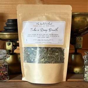 Kraft pouch of Take a Deep Breath loose leaf infusion by The Herbal Cabinet, its window showing dried green leaves, beside brass scales and glass jars of herbs